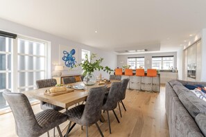Eine moderne Küche mit einer zentralen Kochinsel, ein Essbereich mit einem Holztisch und grau gepolsterten Stühlen sowie ein graues Sofa im Wohnbereich.