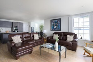 A modern living room with two brown leather sofas, a glass coffee table, and a view of the outside through large windows. - 3 Bed in Carlyon Bay (oc-b623421) (Carlyon Bay)