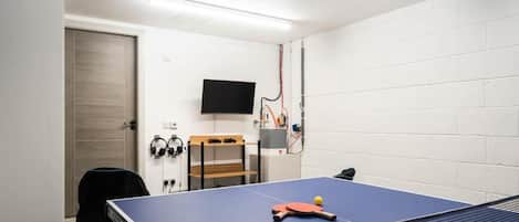 A ping pong table with a ball and paddles, a television mounted on the wall, and a pair of headphones on a shelf.