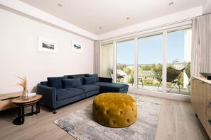 Ein modernes Wohnzimmer mit einer Couch, einem Hocker und Blick ins Freie durch große Fenster.