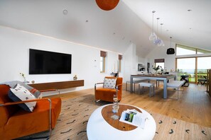 A modern living room with a flat-screen TV, orange sofa, and a dining area with a table and chairs. - 3 Bed in St. Eval (oc-b584290) (Saint Eval)