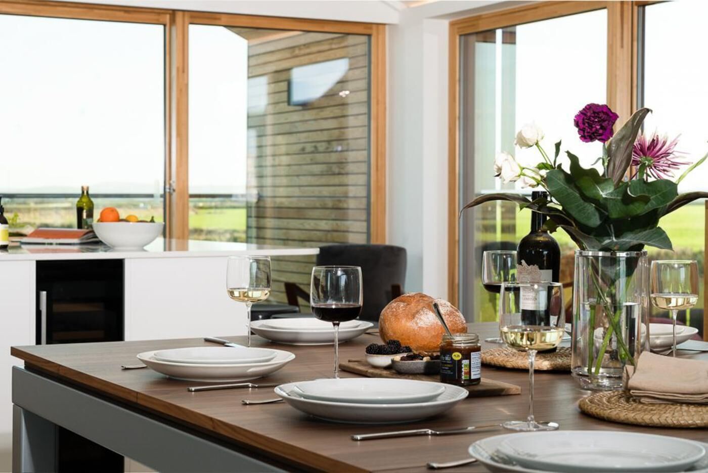 Une table à manger dressée avec des assiettes, des verres, un pain et une bouteille de vin, offrant une vue sur l’extérieur grâce à de grandes fenêtres.