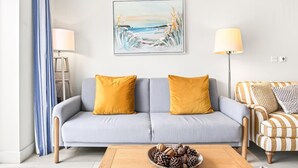 A living room with a sofa, two lamps, a painting, and a coffee table with a pinecone bowl. - 2 Bed in Watergate Bay (oc-b598859) (Tregurrian)