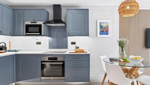 A modern kitchen with grey cabinets, a built-in oven, and a dining area with a glass table and white chairs. - 2 Bed in Watergate Bay (oc-b598859) (Tregurrian)