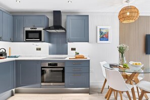 Eine moderne Küche mit grauen Schränken, einem eingebauten Backofen und einem Essbereich mit einem Glastisch und weißen Stühlen.