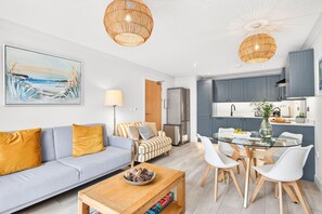 A modern living room with a sofa, coffee table, and dining area with chairs. - 2 Bed in Watergate Bay (oc-b598859) (Tregurrian)