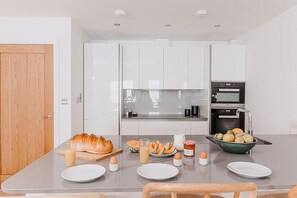 A modern kitchen with white cabinets, a wooden door, and a dining table set with plates, eggs, and a croissant. - 3 Bed in Cawsand (oc-b588795) (Kingsand)