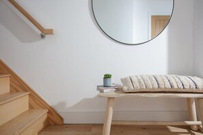 Een houten trap, een ronde spiegel, een kleine plant en een zachte bank tegen een witte muur.