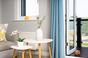 A modern living room with a sofa, a small round table, a potted orchid, and a vase with a plant. There is a window with a view of the outdoors. - 3 Bed in Mawgan Porth (oc-b584450) (Mawgan Porth)