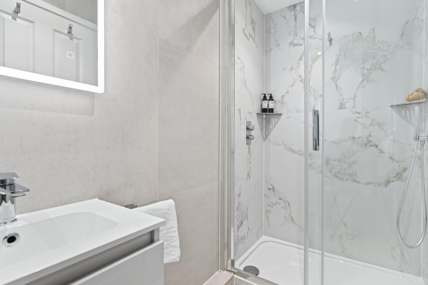Un baño moderno con ducha de vidrio, lavamanos blanco y pared con mosaicos de mármol.