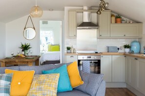 A compact kitchen with a built-in oven, a dining area with a table set for two, and a cozy seating area with a sofa. - 2 Bed in Whitsand Bay (oc-b584334) (Freathy)