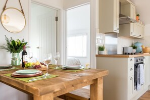 A dining area with a wooden table set for a meal, a bottle of wine, and a vase with flowers. - 2 Bed in Whitsand Bay (oc-b584334) (Freathy)