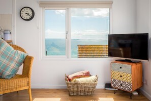 A room with a television, wicker furniture, a wicker basket, and a view of the sea through a window. - 2 Bed in Whitsand Bay (oc-b584334) (Freathy)