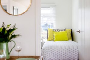 A bedroom with a bed, a mirror, a vase with flowers, and a window with blinds. - 2 Bed in Whitsand Bay (oc-b584334) (Freathy)