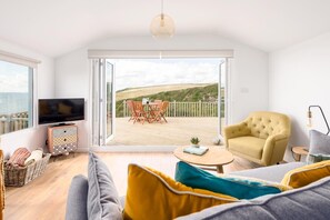A modern living room with a large sliding glass door leading to an outdoor deck, a television, a sofa, and a yellow armchair. - 2 Bed in Whitsand Bay (oc-b584334) (Freathy)