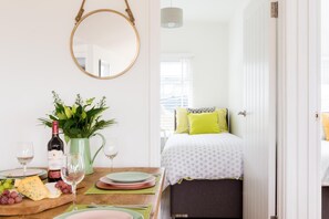A room with a bed, a dining table with plates, glasses, and a bottle of wine, and a hanging mirror. - 2 Bed in Whitsand Bay (oc-b584334) (Freathy)