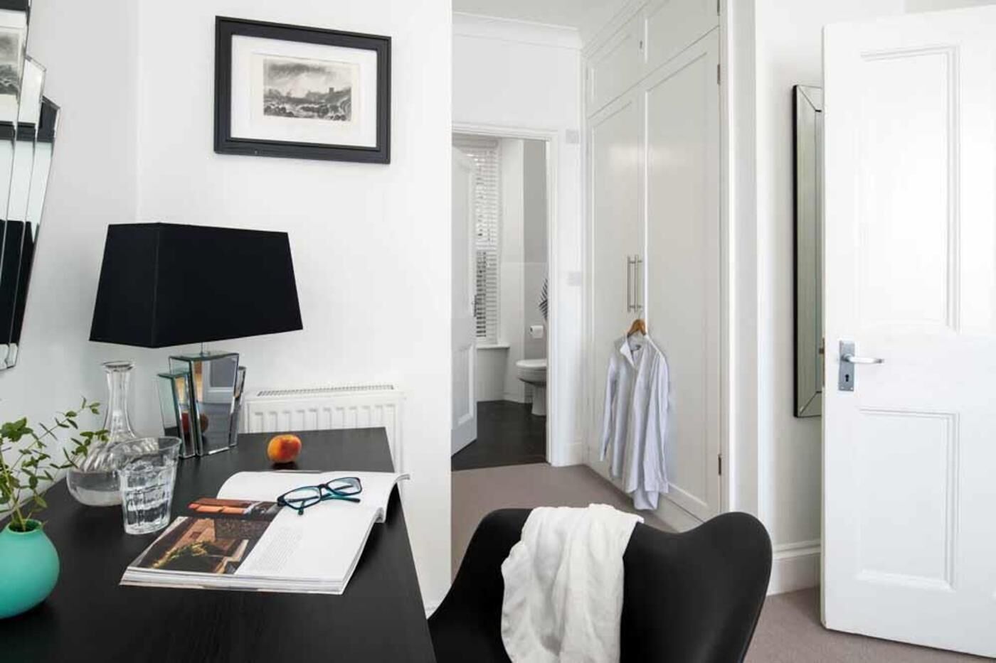 Ein Hotelzimmer mit Schreibtisch, einem gerahmten Bild, einem Heizkörper, einem Spiegel, einer Zimmerpflanze und einer Tür zu einem Badezimmer.