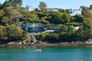 A coastal property with a modern house, a boat on the water, and a hillside with dense vegetation. - 4 Bed in Fowey (oc-b584335) (Fowey)