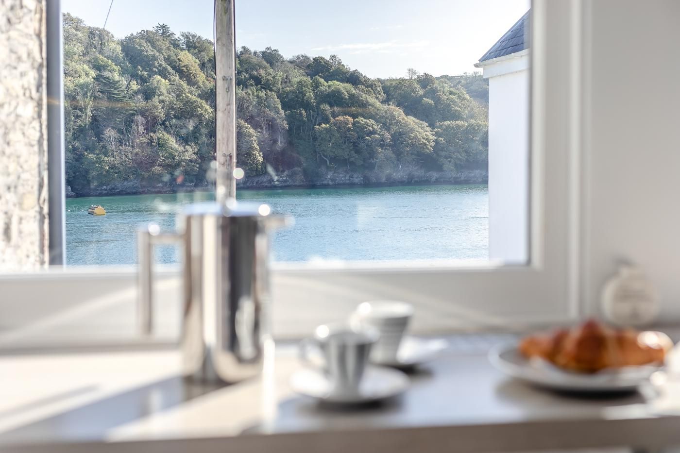 Une chambre avec vue sur un plan d’eau et une colline boisée. Sur le rebord de la fenêtre, on trouve une machine à café, une tasse et une assiette avec de la nourriture.