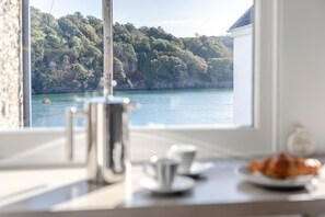 Une chambre avec vue sur un plan d’eau et une colline boisée. Sur le rebord de la fenêtre, on trouve une machine à café, une tasse et une assiette avec de la nourriture.