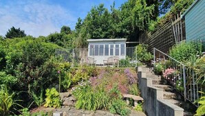 A garden with a wooden deck, a stone wall, and a small building with a metal fence. - 4 Bed in Fowey (oc-b584335) (Fowey)