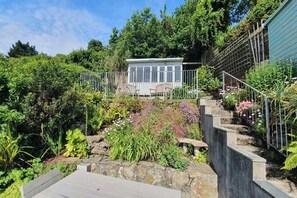 A garden with a wooden deck, a stone wall, and a small building with a metal fence. - 4 Bed in Fowey (oc-b584335) (Fowey)