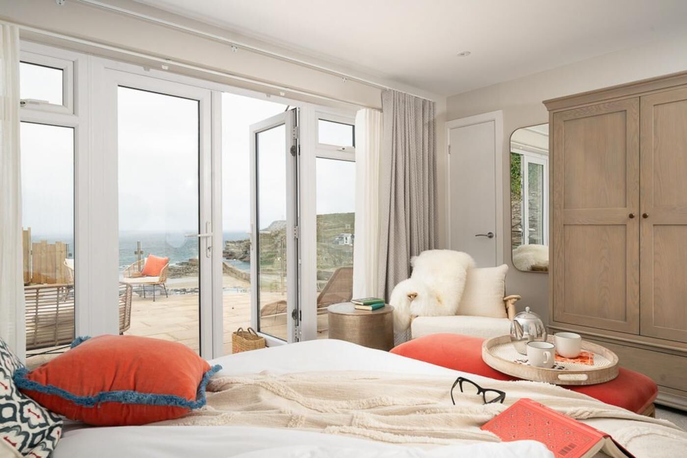 A hotel room with a bed, a chair, a side table, and a wardrobe. There is a view of the beach and the sea through the open door.