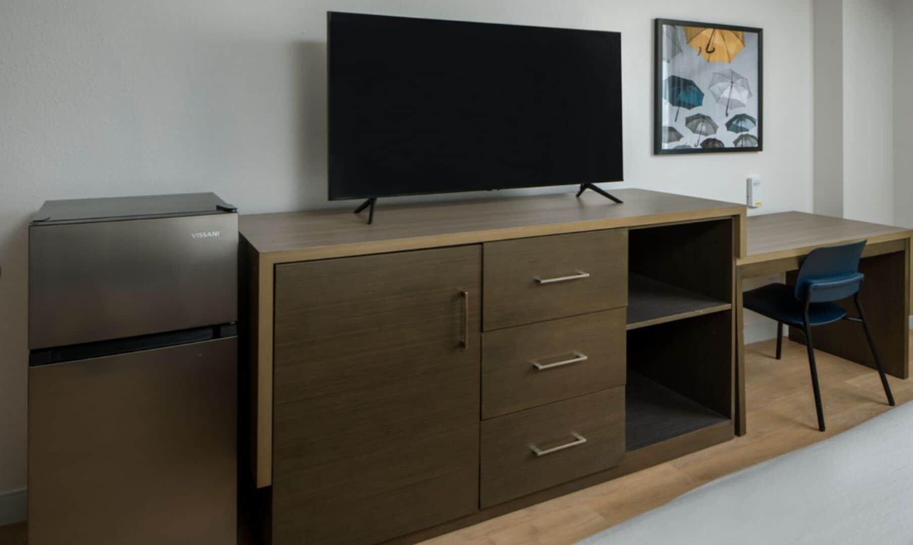 A compact room with a desk, a flat-screen TV, a VISSANI refrigerator, and a chair.