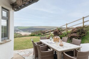 An outdoor patio with wicker furniture, a table set with a vase, and a scenic view of hills and a river. - 4 Bed in Mawgan Porth (oc-b650080) (Mawgan Porth)