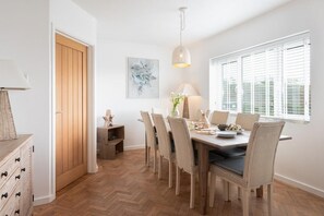A dining area with a wooden table, wicker chairs, a hanging light fixture, and a window with blinds. - 4 Bed in Mawgan Porth (oc-b650080) (Mawgan Porth)