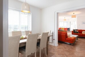 Een eetgedeelte met rieten stoelen en een tafel, een feloranje bank in de woonkamer, en uitzicht door een open deur.