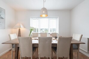 A dining area with a wooden table, six wicker chairs, a hanging light fixture, and a window with blinds. - 4 Bed in Mawgan Porth (oc-b650080) (Mawgan Porth)