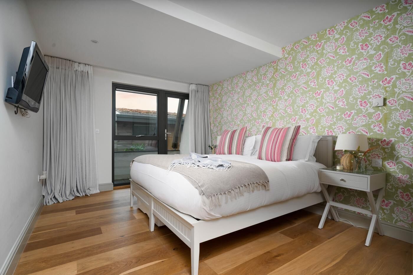 Een slaapkamer met een bed, gordijnen, een televisie, een nachtkastje en behang met een patroon.