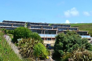Ein modernes, mehrstöckiges Gebäude mit mehreren Balkonen und großen Fenstern, umgeben von üppiger Vegetation und blauem Himmel.