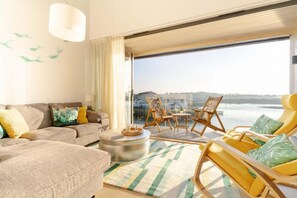 A modern living room with a sofa, armchairs, a coffee table, and a view of a body of water through large windows. - 3 Bed in Hayle (oc-b635644) (Hele)