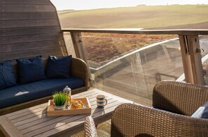 Ein Balkon mit einer Rattan-Couch und -Stühlen, einem Holztisch mit einer Essschale und einer Tasse, und Blick auf eine hügelige Landschaft.