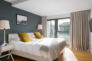 A modern hotel room with a large bed, two bedside lamps, a framed picture on the wall, and a view through a large window. - 2 Bed in Watergate Bay (oc-b584354) (Tregurrian)