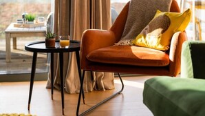 A modern living room with a comfortable orange chair, a small round table with a plant and a glass of orange juice, and a view of the outdoors. - 2 Bed in Watergate Bay (oc-b584354) (Tregurrian)