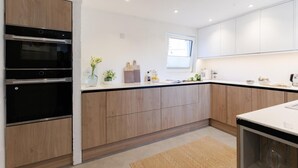A modern kitchen with built-in ovens, wooden cabinets, and a window providing natural light. - 3 Bed in Downderry (oc-b652220) (Downderry)