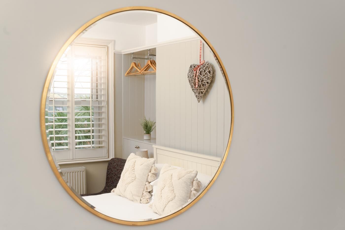 Un dormitorio con un espejo redondo, ropa de cama blanca en una cama, un tocador, una ventana con persianas y un adorno con forma de corazón.
