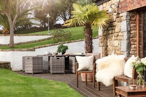 An outdoor patio with wicker furniture, a stone wall, and a palm tree. - 5 Bed in Holywell Bay (oc-b614951) (Cubert)