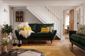 Ein Wohnzimmer mit einem grünen Sofa, einem Couchtisch mit Weingläsern und einem Buch, und einer Treppe im Hintergrund.