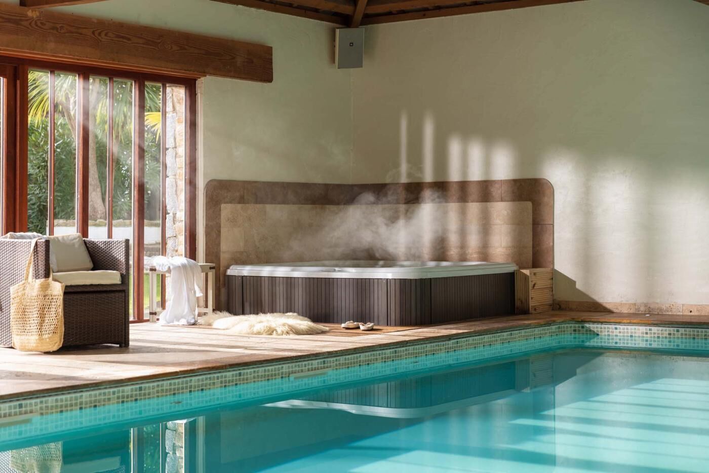 Un bain à remous dont s’échappe de la vapeur, une piscine à l’eau d’un bleu limpide et un canapé en osier agrémenté de coussins.