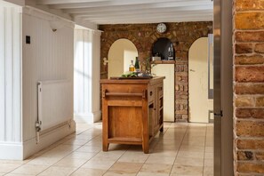 A kitchen with a wooden table, a white radiator, and a brick wall with a clock. - 5 Bed in Holywell Bay (oc-b614951) (Cubert)