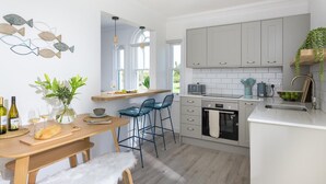 A modern kitchen with a wooden table, bar stools, a built-in oven, and a window overlooking the garden. - 2 Bed in Fistral (oc-b625209) (Newquay)