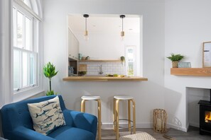 Ein modernes Wohnzimmer mit einem blauen Sofa, zwei Hocker, einem Kamin und einer durch einen Rundbogen sichtbaren Küche.