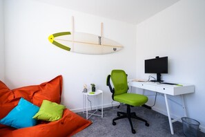 A room with a white desk, a green office chair, a black monitor, a surfboard decoration, a small side table, and a colorful orange couch. - 3 Bed in Fistral (oc-b595429) (Newquay)
