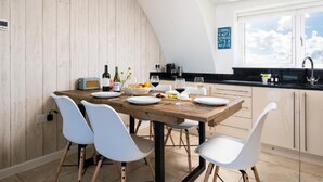 A dining area with a wooden table, white chairs, and a kitchen in the background. - 3 Bed in Fistral (oc-b595429) (Newquay)