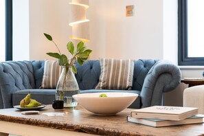 A modern living room with a blue sofa, a glass vase with green leaves, a bowl with a lime, and a book on a wooden table. - 5 Bed in Carbis Bay (oc-b623959) (Carbis Bay)