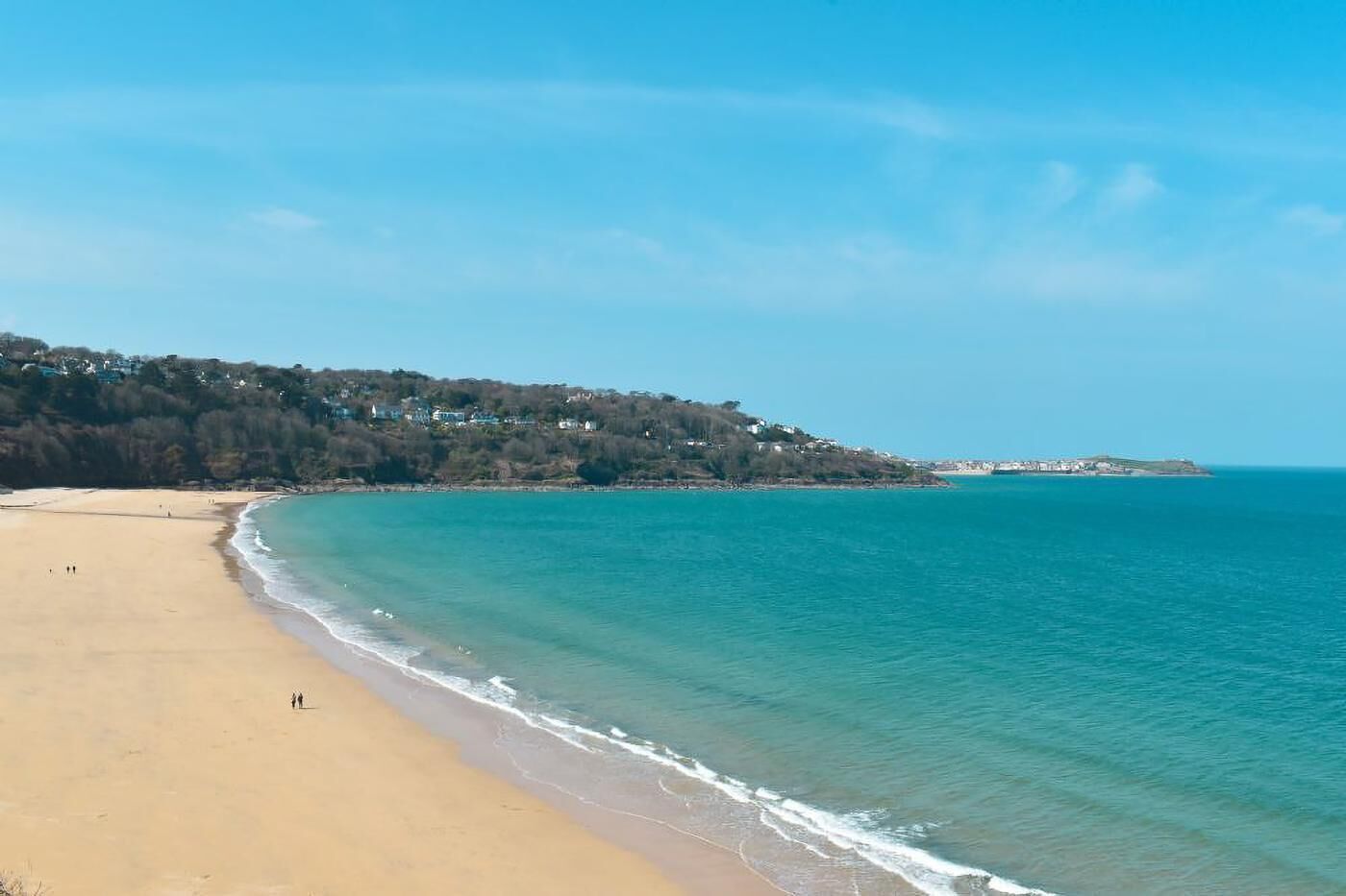 A beach with clear turquoise water, a sandy shore, and a forested hill in the background.